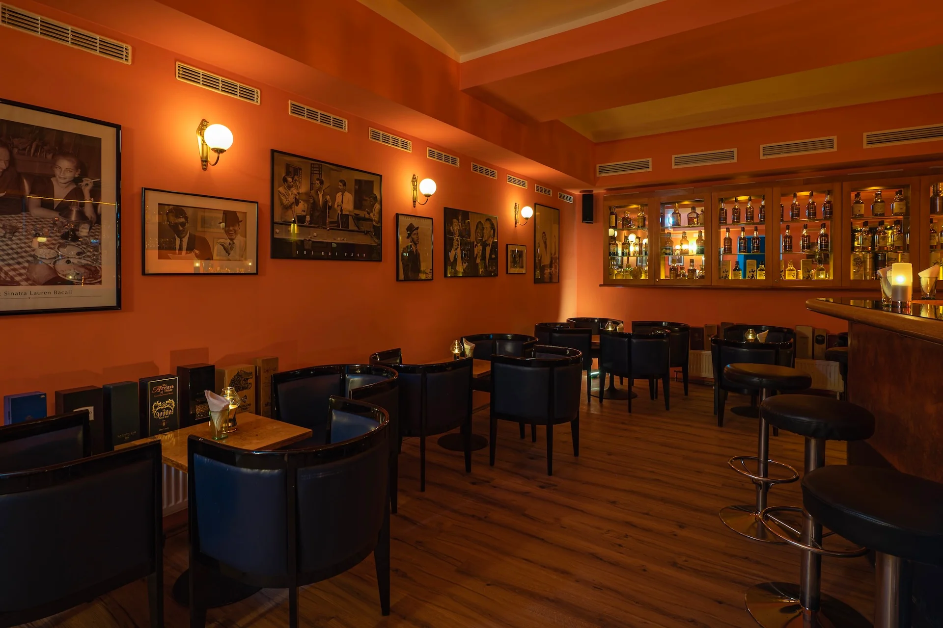 Eine gemütliche Bar mit orangefarbenen Wänden, schwarzen Stühlen, Holztischen, gerahmten Fotos und einem gut beleuchteten Regal hinter der Theke, in dem Flaschen ausgestellt sind. Der Raum verfügt über eine warme Beleuchtung und einen Holzboden, was eine einladende Atmosphäre schafft.