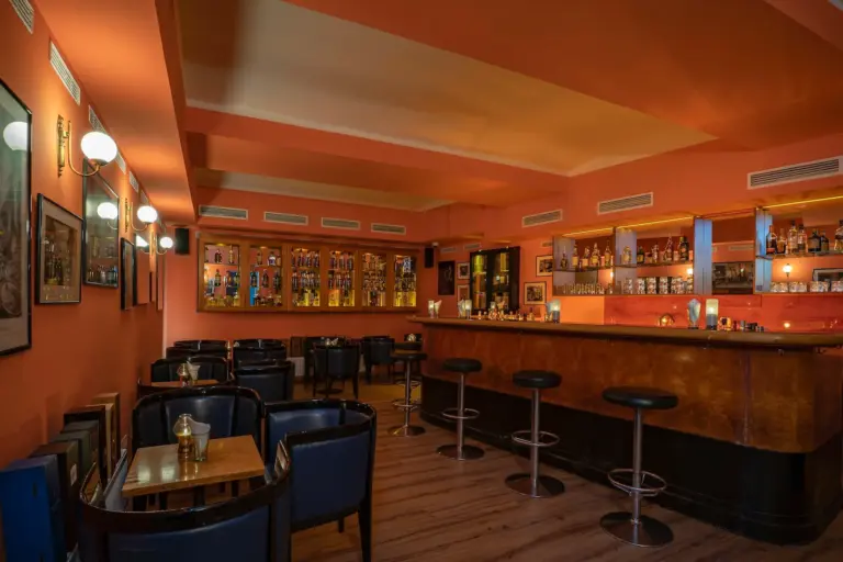 Castillo's The Club Eine gemütliche Bar mit korallenfarbenen Wänden, schwarzen Barhockern an der Theke, dunklen Ledersesseln, kleinen Holztischen und Regalen voller Flaschen hinter der Theke, die von Wandleuchten und Deckenlampen in warmes Licht getaucht sind.