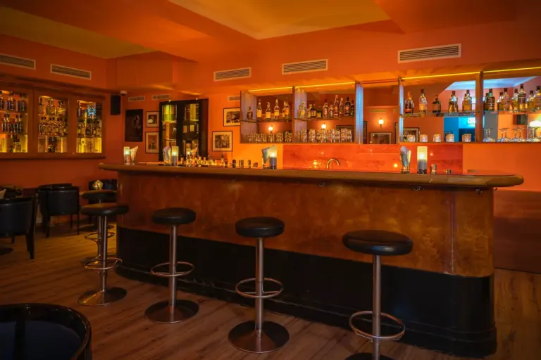 Eine warm beleuchtete Bar mit Regalen voller Flaschen, einer polierten Holztheke, hohen Hockern und kerzenbeleuchteten Tischen, die eine gemütliche und einladende Atmosphäre schaffen.