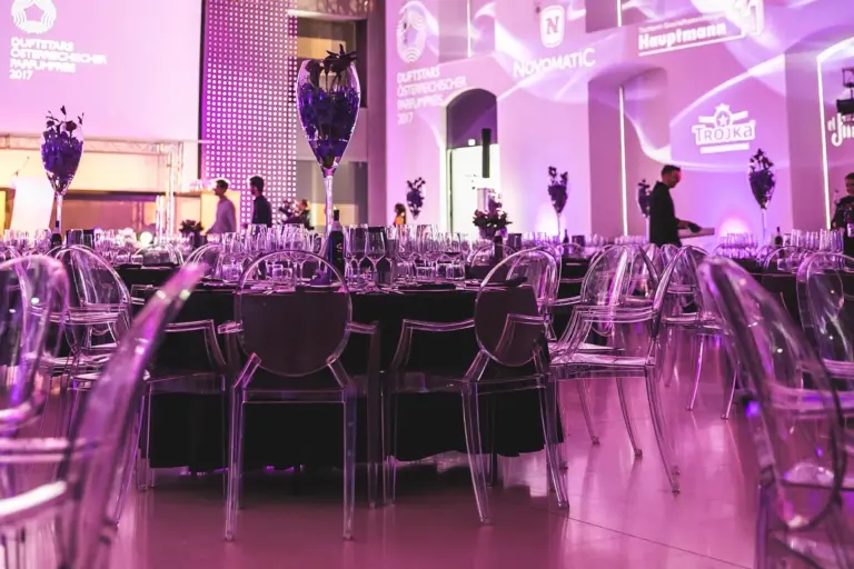 Cateringkultur Ein Bankettsaal mit runden Tischen, die für eine Veranstaltung gedeckt sind, mit durchsichtigen Stühlen, eleganten Tischdecken und violetter Beleuchtung. Im Hintergrund stehen Menschen an beleuchteten Wänden mit projizierten Logos.