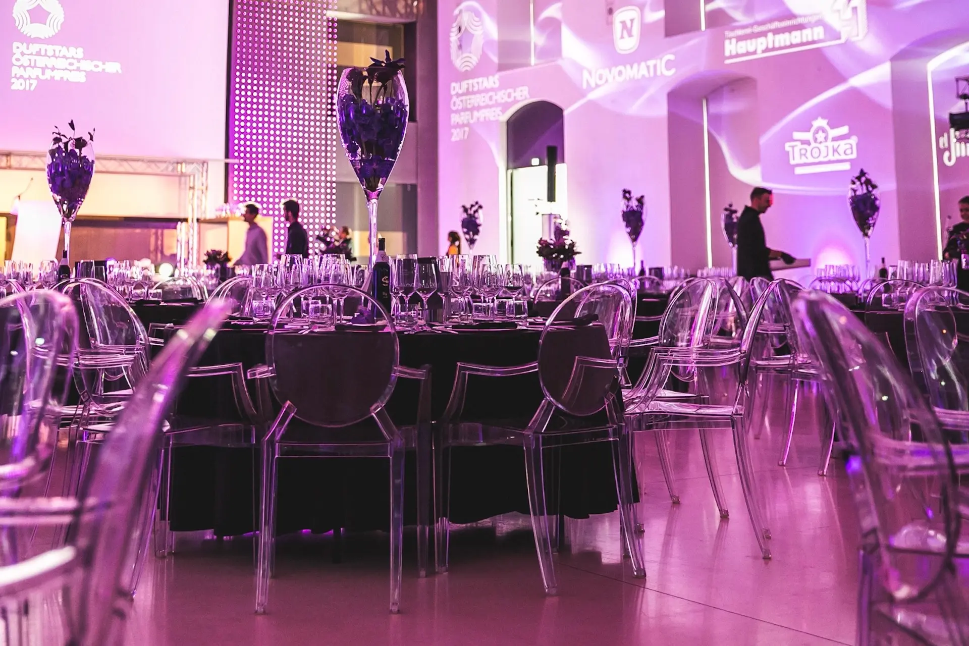 Cateringkultur Ein Bankettsaal mit runden Tischen, die für eine Veranstaltung gedeckt sind, mit durchsichtigen Stühlen, eleganten Tischdecken und violetter Beleuchtung. Im Hintergrund stehen Menschen an beleuchteten Wänden mit projizierten Logos.