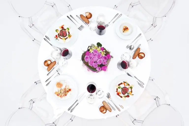 Ein runder weißer Tisch für vier Personen mit Gourmetgeschirr, Rotweingläsern, Brötchen, Silberbesteck und einem violetten Blumenschmuck. Klare Stühle umgeben den Tisch, von oben gesehen.