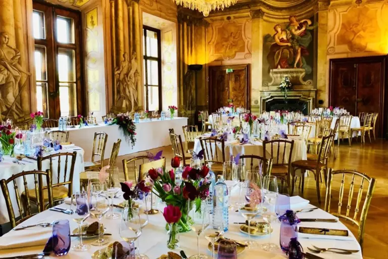 Eleganter Bankettsaal mit verschnörkelten Wänden und großen Fenstern, dekoriert mit runden Tischen für eine formelle Veranstaltung. Die Tische sind mit Blumenkränzen, Weingläsern und goldenen Stühlen geschmückt, im Hintergrund ein großer Kamin.