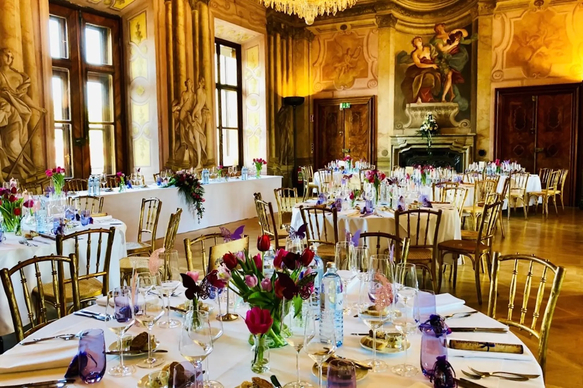 Eleganter Bankettsaal mit verschnörkelten Wänden und großen Fenstern, dekoriert mit runden Tischen für eine formelle Veranstaltung. Die Tische sind mit Blumenkränzen, Weingläsern und goldenen Stühlen geschmückt, im Hintergrund ein großer Kamin.