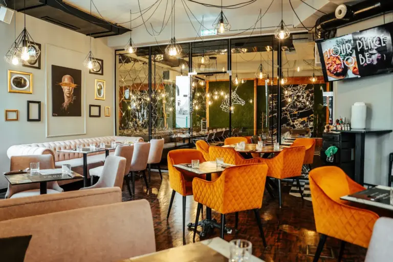 Modernes Restaurantinterieur mit gelben und beigen Stühlen, Marmortischen, hängenden Leuchten, gerahmten Kunstwerken an den Wänden und einem Fernseher in der Ecke. Der Raum bietet eine gemütliche, stilvolle und einladende Atmosphäre.