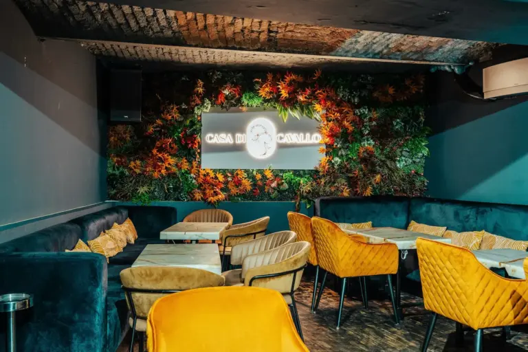 Ein stilvolles Restaurant mit Samtstühlen - manche senfgelb, manche dunkelblau - um Marmortische herum. An der Rückwand hängen grüne Blätter und ein Neonschild mit der Aufschrift Casa do Cavalo.