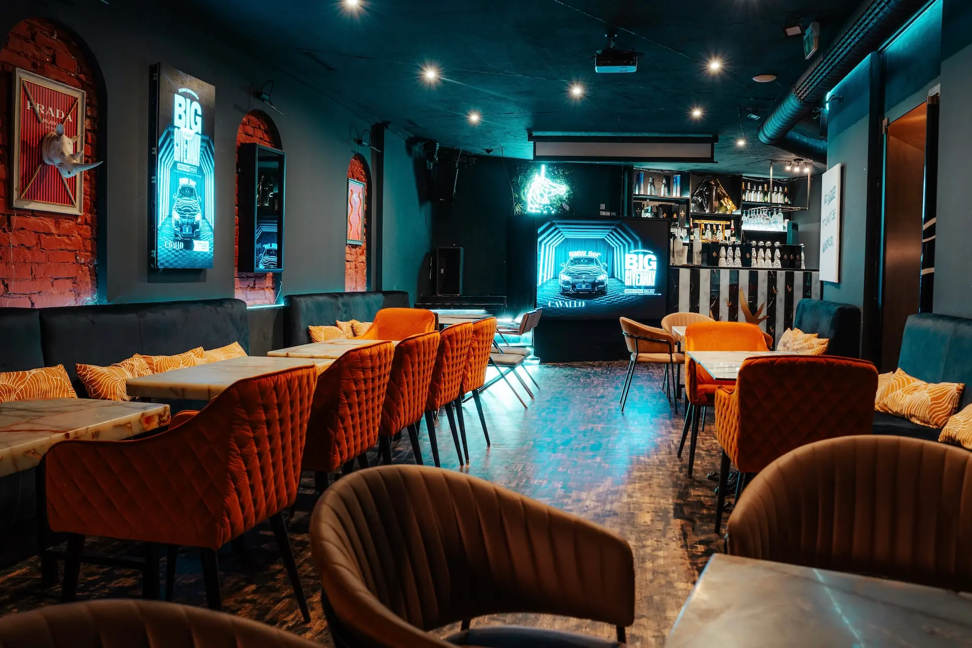 Cavallo Eine moderne Lounge-Bar mit orangefarbenen und braunen Stühlen, kleinen Tischen, Leuchtreklamen, Postern und einem gut beleuchteten Barbereich im Hintergrund, der eine gemütliche und einladende Atmosphäre schafft.