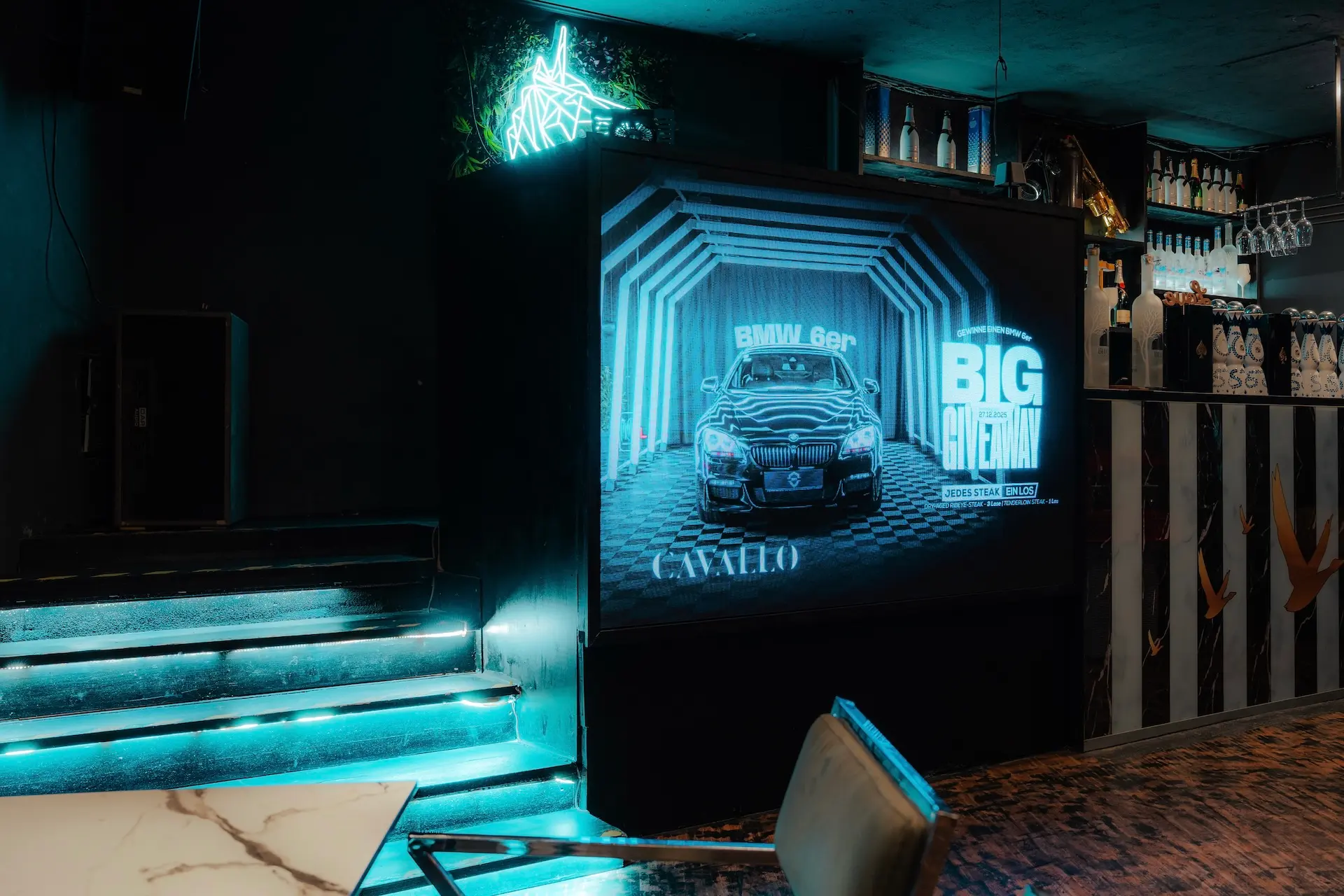 In einem dunklen Raum mit Neonbeleuchtung ist ein großer Bildschirm zu sehen, auf dem eine BMW-Werbung für ein GROSSES GEWINNSPIEL läuft. Auch Regale mit Flaschen und ein Marmortisch sind zu sehen.
