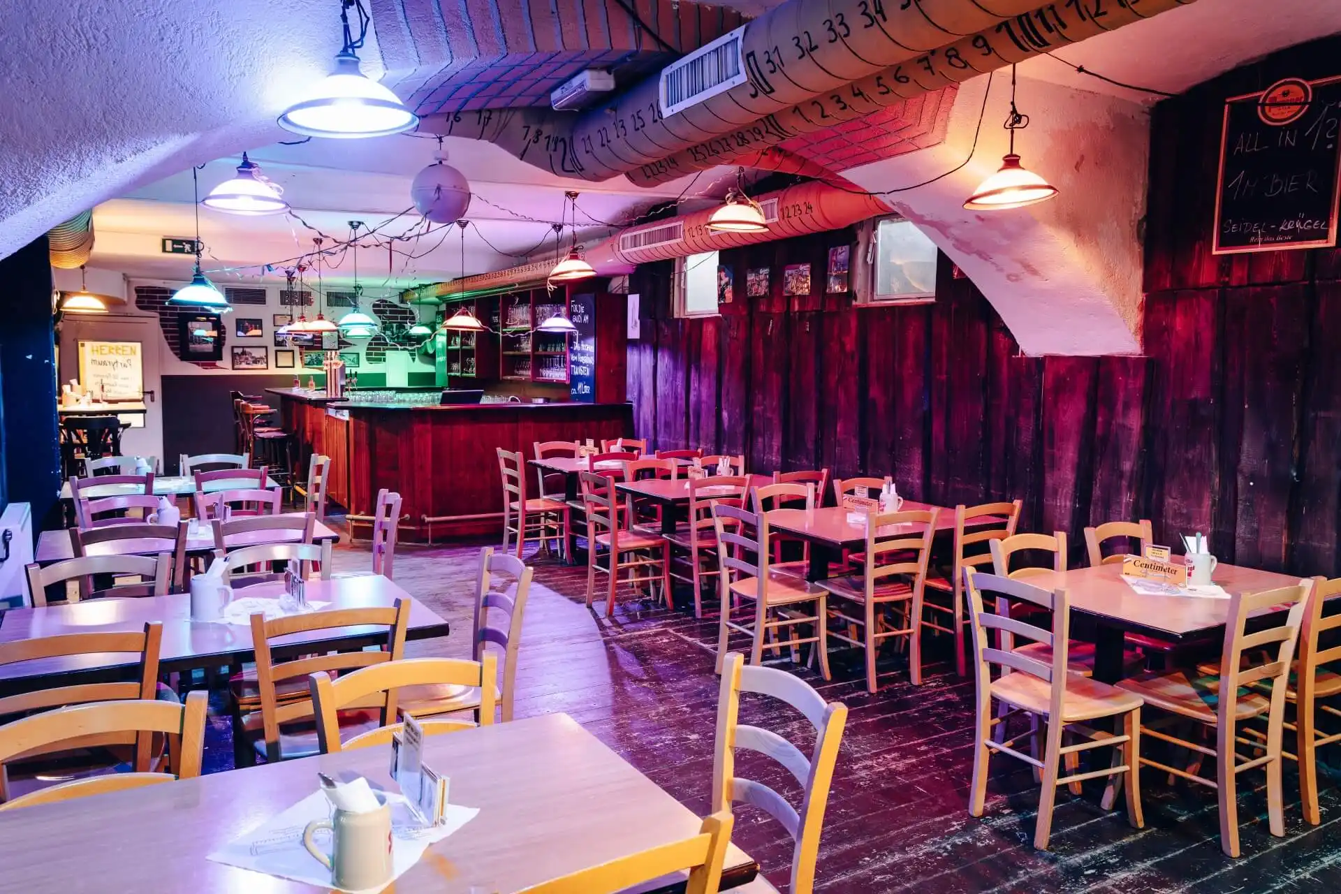 Ein gemütliches, schummriges Restaurant mit Holztischen und -stühlen, einer gewölbten Decke und bunten Hängelampen. Der Barbereich befindet sich im hinteren Teil, und die Wände sind mit dekorativen Schriftzügen und kleinen gerahmten Bildern geschmückt.