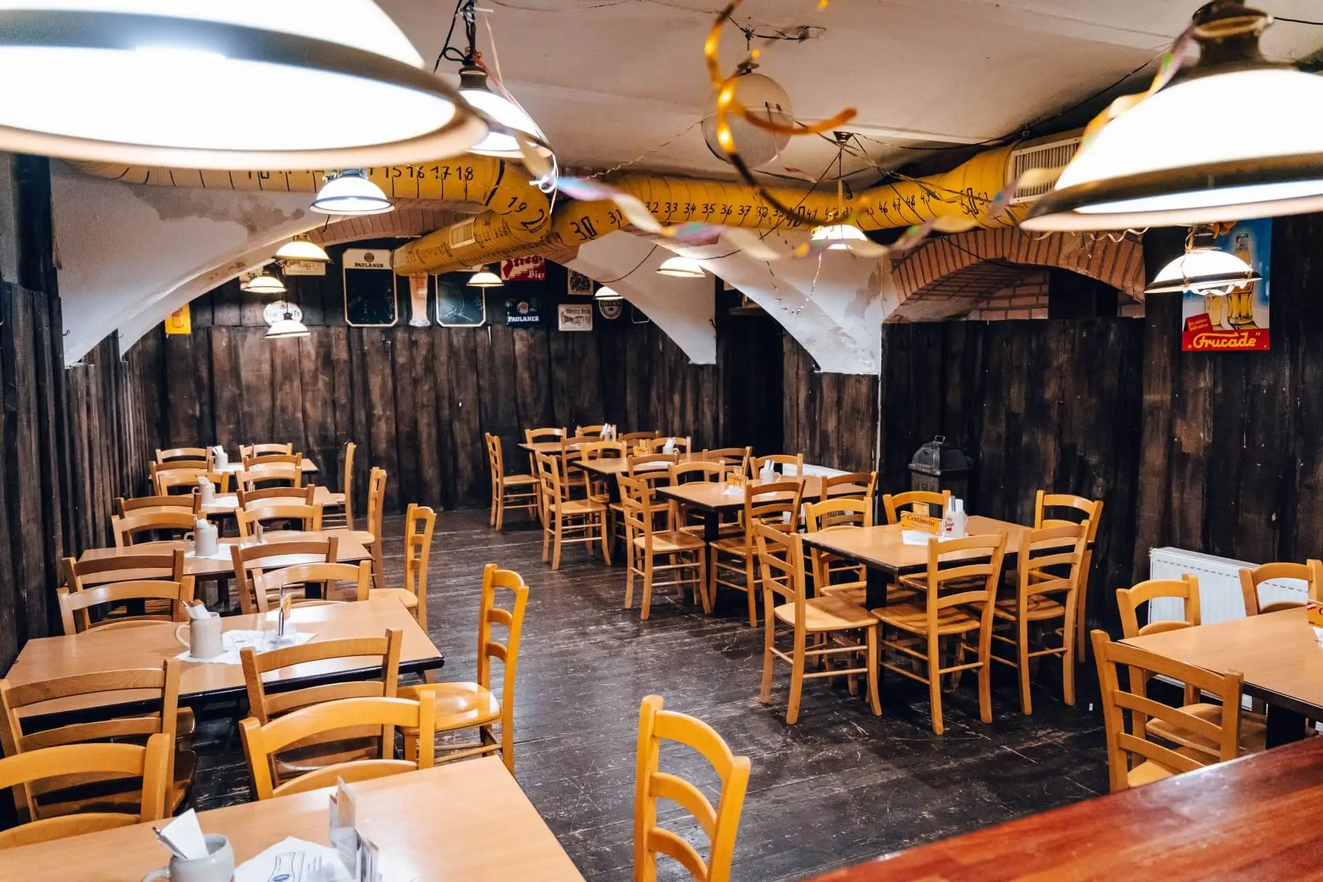 Ein gemütlicher Restaurantbereich mit leeren Holztischen und -stühlen, warmer Deckenbeleuchtung, rustikalen Wänden aus dunklem Holz und von der Decke hängenden Dekorationsgegenständen.