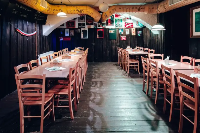 Ein rustikaler Restaurantbereich mit Holzfußboden und langen Tischen mit Holzstühlen. Bunte Fotos und Plakate schmücken die dunklen Wände, und an der Decke verläuft ein gelber Lüftungsschacht.