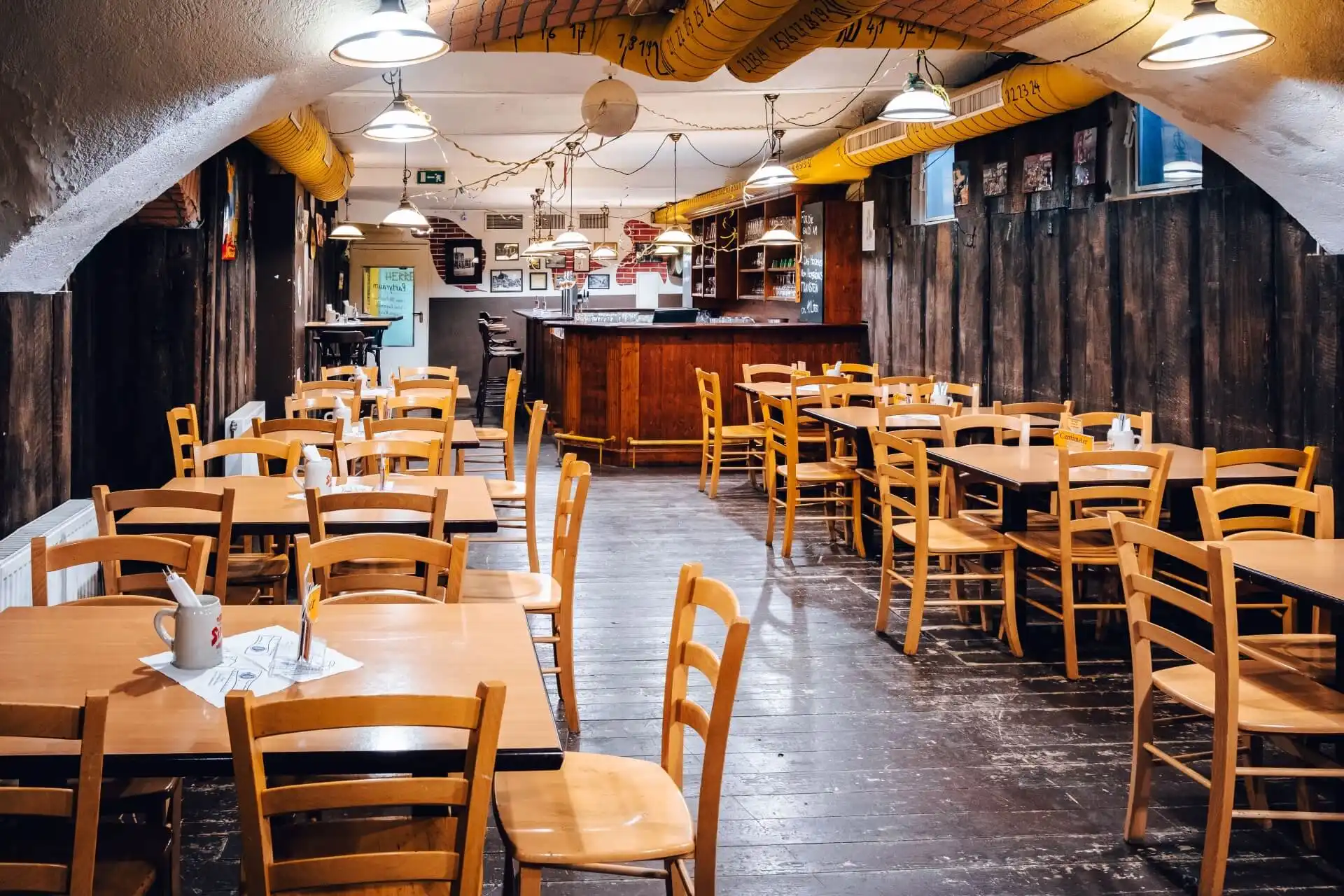 Ein gemütliches, rustikales Restaurant mit Holztischen und -stühlen, gelben Luftschächten an der gewölbten Decke, warmer Beleuchtung und einem hölzernen Bartresen im hinteren Bereich. Der Essbereich wirkt leer und einladend.