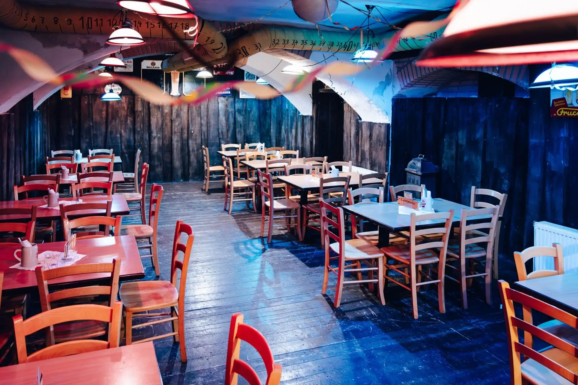 Ein farbenfrohes, leeres Restaurant mit Holztischen und -stühlen, blau getönter Beleuchtung und von der Decke hängenden Luftschlangen. Die Wände haben einen rustikalen Look, und über dem Sitzbereich ist Zahlenschmuck drapiert.