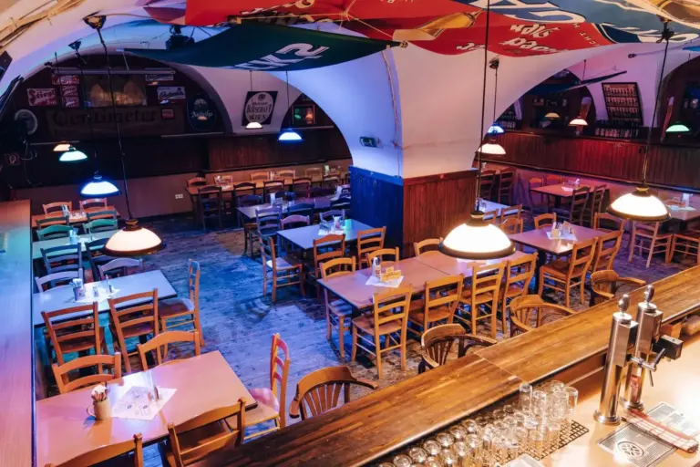 Ein gemütliches, leeres Restaurant mit Holztischen und -stühlen, bunten Hängelampen und einer Bar mit Gläsern und Bierhähnen im Vordergrund. Die Decke ist gewölbt und mit verschiedenen Bannern geschmückt.