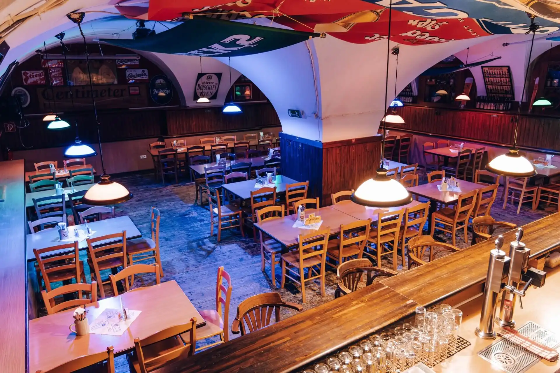 Ein gemütliches, leeres Restaurant mit Holztischen und -stühlen, bunten Hängelampen und dekorativen Schirmen an der Decke. Im Vordergrund ist eine hölzerne Bar mit Gläsern und Bierhähnen zu sehen.
