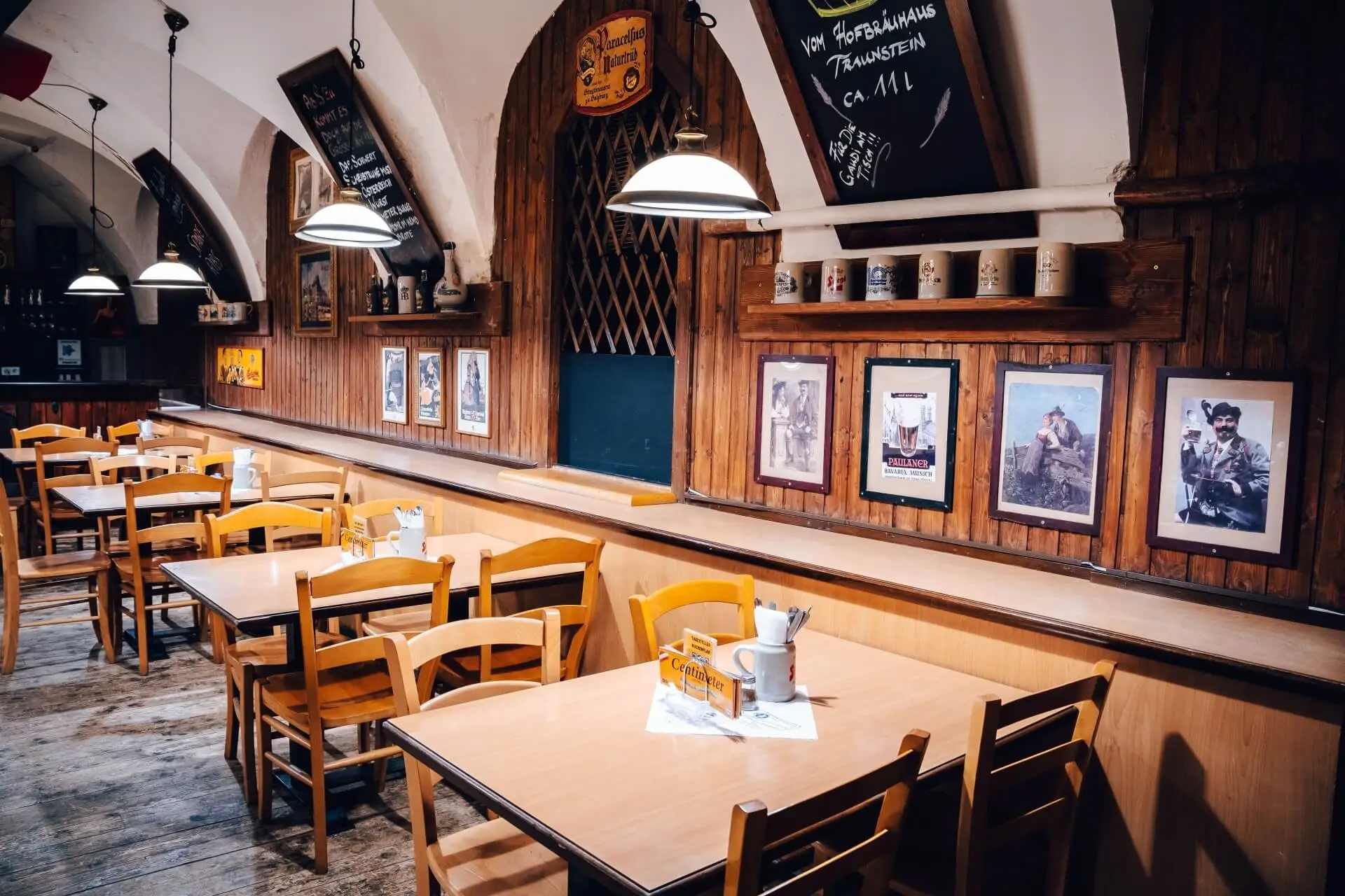 Ein gemütliches, leeres Restaurant mit Holztischen und -stühlen, warmer Beleuchtung und gerahmten Bildern an holzgetäfelten Wänden. Die Speisekarten hängen an einer Kreidetafel und die Tassen stehen auf einem Regal über dem Sitzbereich.