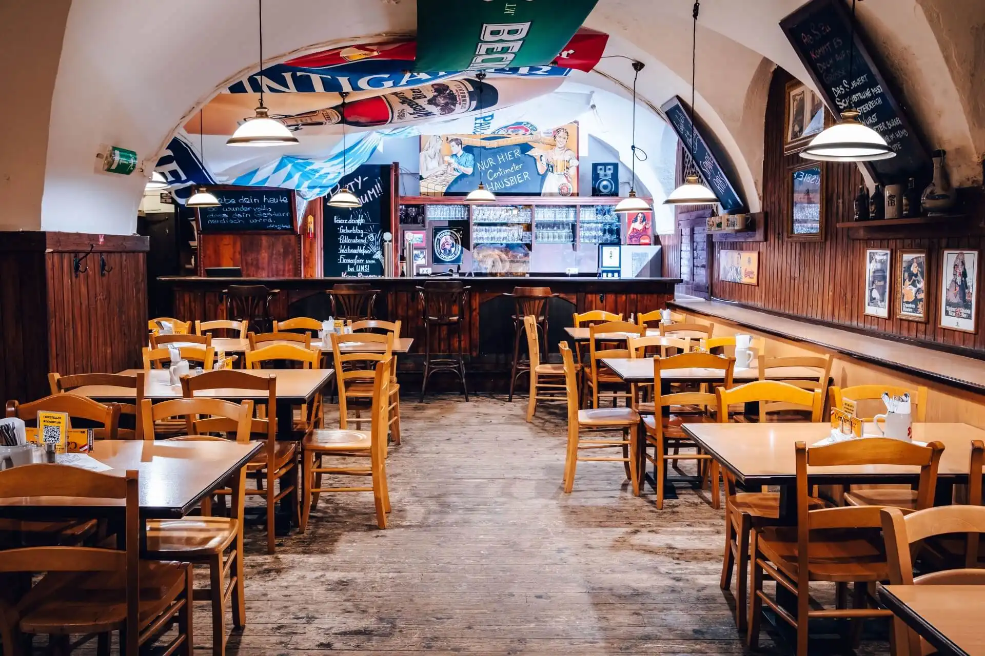 Eine gemütliche Kneipe mit Holztischen und -stühlen, von der gewölbten Decke hängenden Sportbannern, Speisekarten auf Kreidetafeln und einem mit Flaschen und Gläsern bestückten Barbereich. Der Raum ist leer und warm beleuchtet.