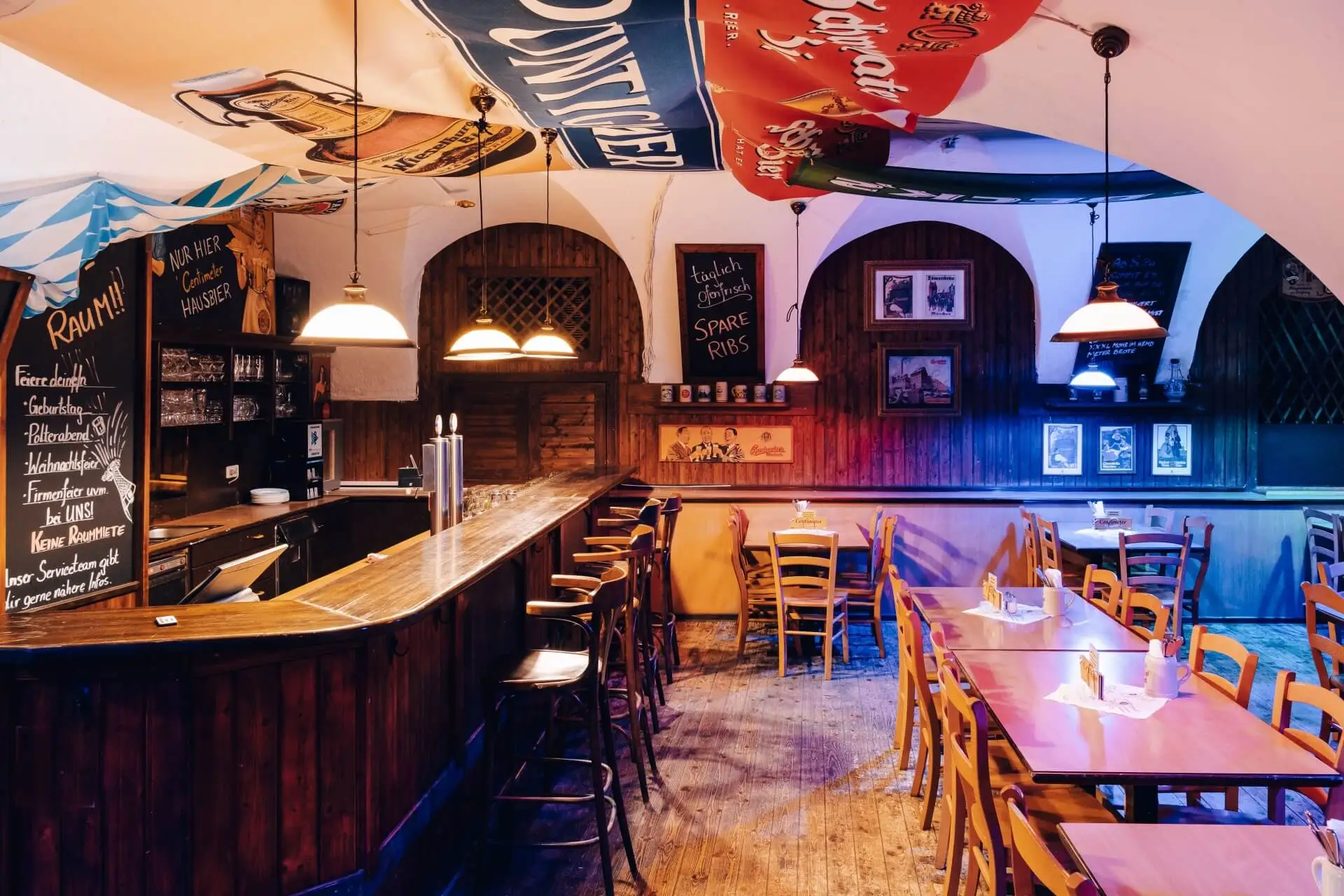 Eine gemütliche, leere Kneipe mit Holztischen und -stühlen, einer langen Holzbar, hängenden Pendelleuchten, bunten Bannern an der Decke und gerahmten Bildern an getäfelten Wänden, die eine warme, einladende Atmosphäre schaffen.