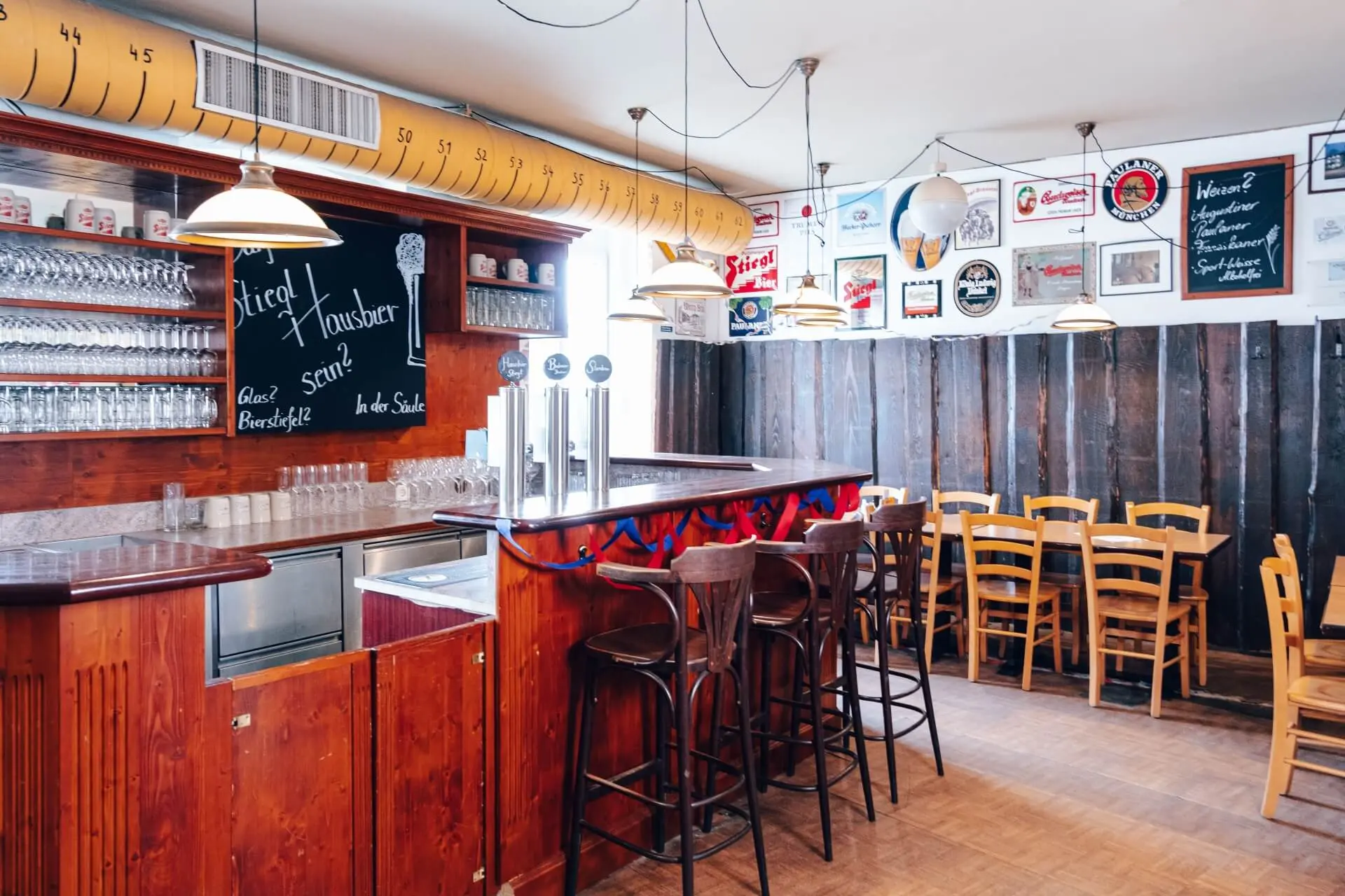 Gemütliche Bar mit einem Holztresen, hohen Hockern und Tischen mit Stühlen. Gläser stehen in Regalen, Bierzapfhähne auf dem Tresen und alte Bierschilder zieren die weißen Wände über der dunklen Holzvertäfelung. Von der Decke hängen warme, hängende Lampen.