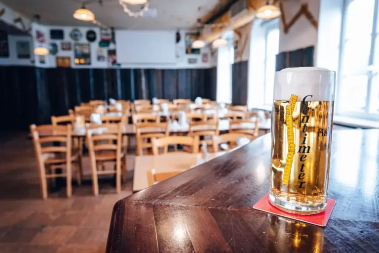 Ein Bierglas mit einem Zentimeterlineal auf dem Tresen; im Hintergrund leere Holzstühle und Tische in einem hellen Restaurant oder einer Bierhalle.