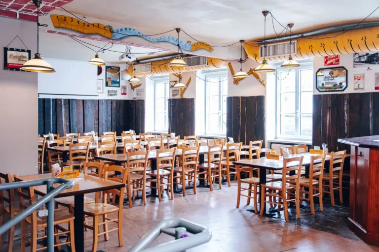 Ein hell erleuchteter Restaurantbereich mit vielen leeren Holztischen und -stühlen, großen Fenstern, die natürliches Licht hereinlassen, und einer skurrilen Einrichtung mit Wandkunst, Hängelampen und Maßbändern an der Decke.