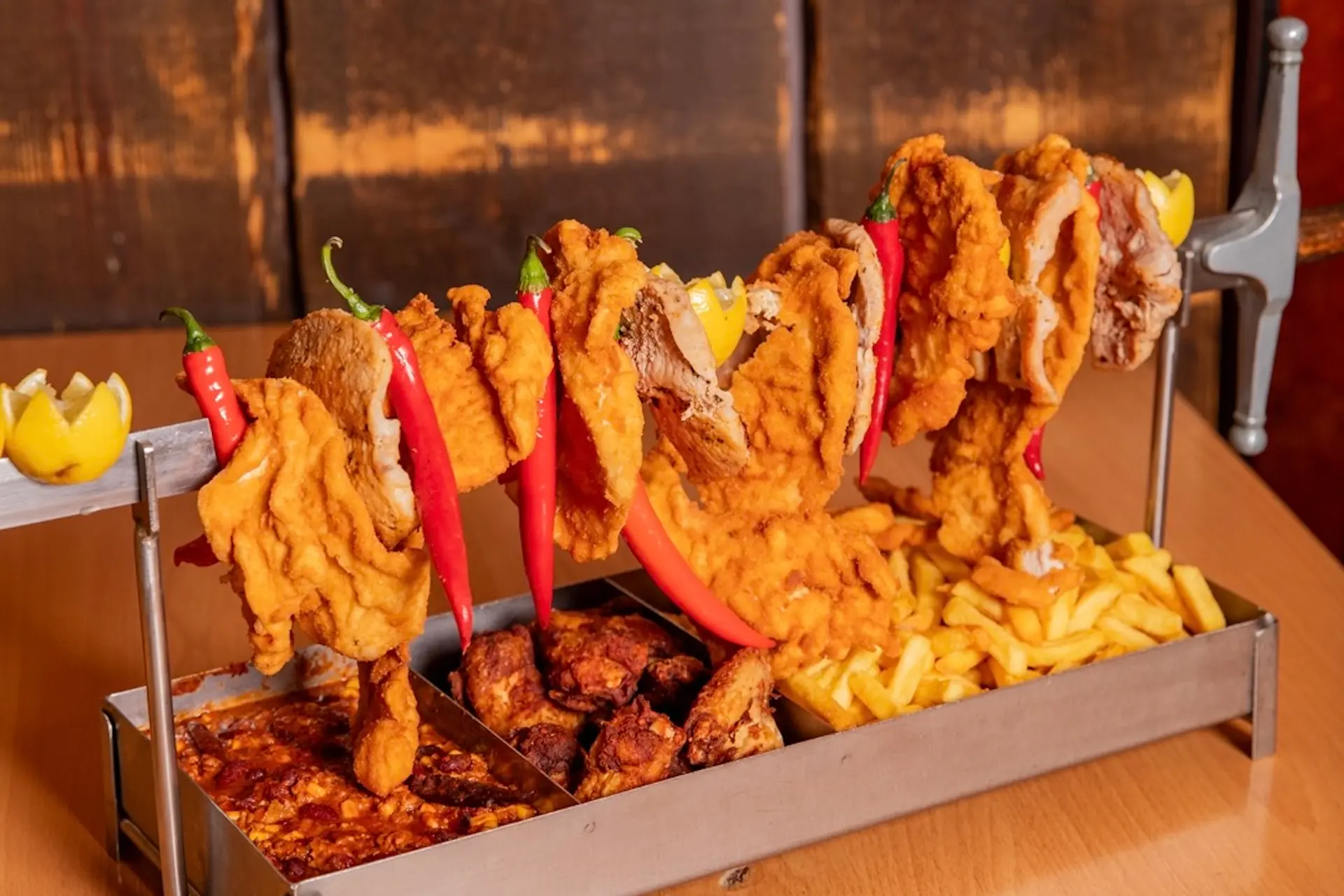 Auf einem Metallgestell liegen panierte Hähnchenteile und rote Chilischoten, darunter befinden sich Pommes frites, scharfe Hähnchenflügel und Baked Beans, die auf einem Holztisch serviert werden.