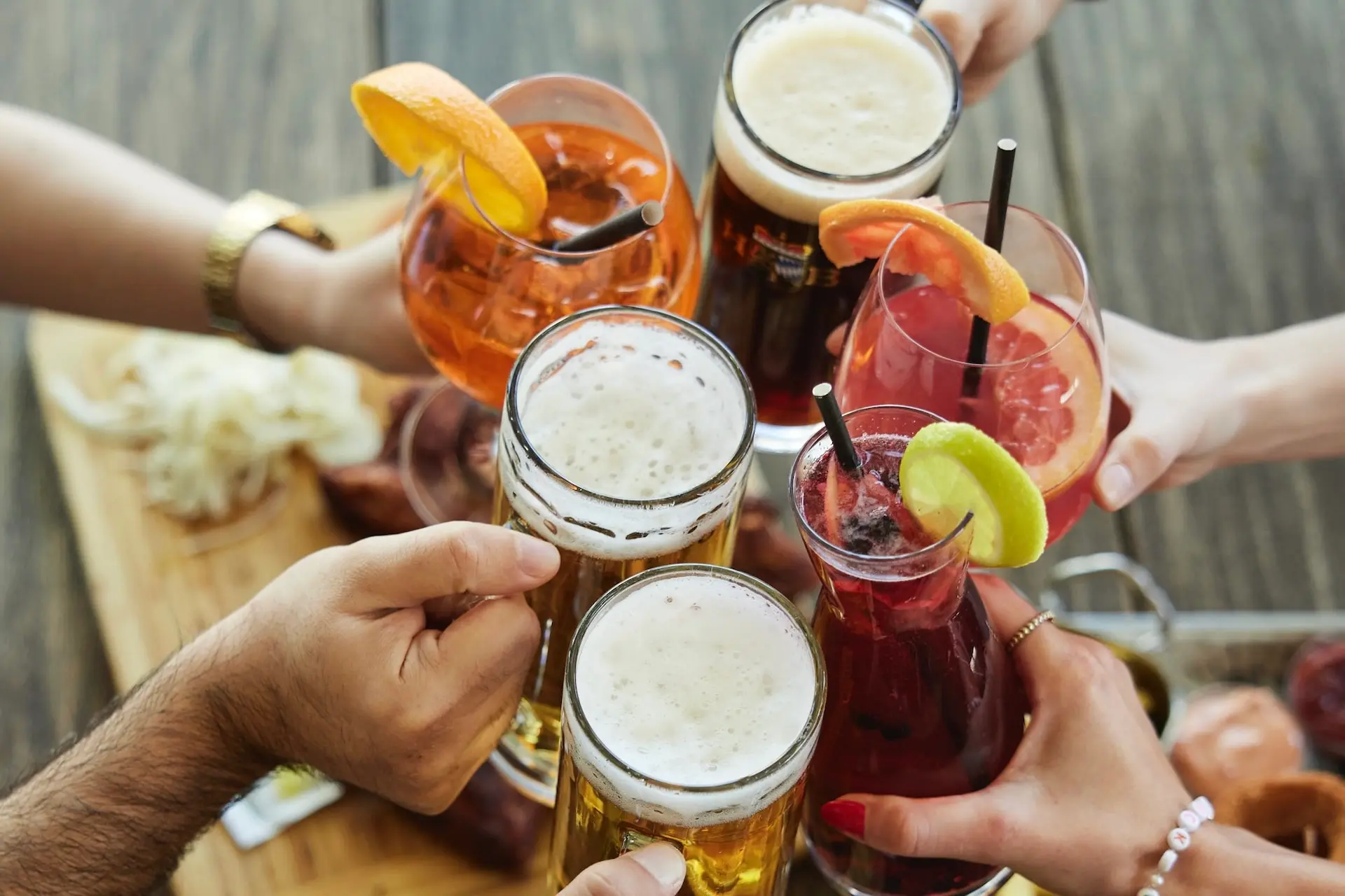 Eine Gruppe von Menschen erhebt verschiedene Getränke, darunter Bierkrüge und bunte, mit Früchten garnierte Cocktails, zu einem Toast auf einen Holztisch mit Essen im Hintergrund.