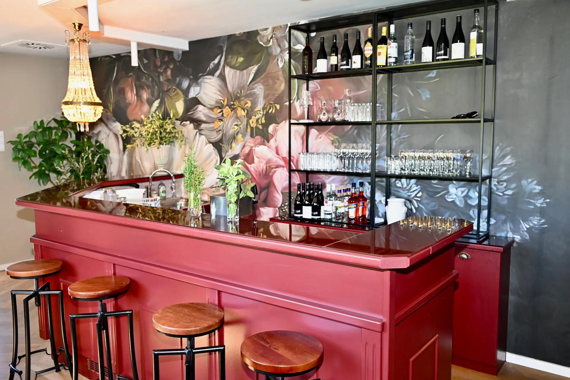 Eine stilvolle Bar mit einer glänzend roten Theke, Holzhockern und Regalen mit Gläsern und Flaschen. Im Hintergrund ist ein großes Blumengemälde zu sehen, und zur Dekoration gibt es Pflanzen und einen Kronleuchter.