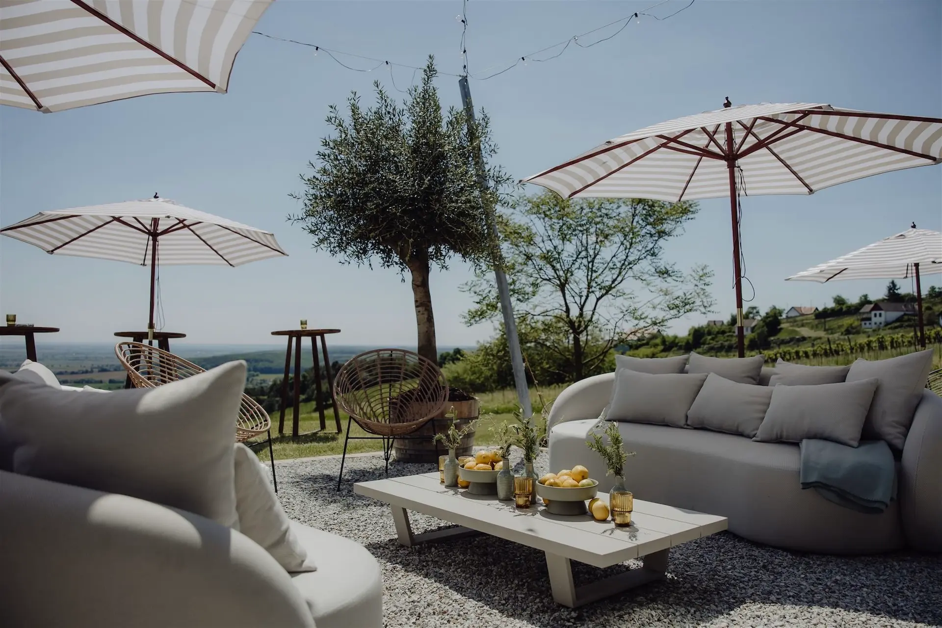 Lounge-Bereich im Freien mit gepolsterten Sofas, einem niedrigen Couchtisch mit Zitronen und Getränken, gestreiften Sonnenschirmen, runden Korbstühlen und einer malerischen Aussicht auf Bäume und eine ferne Landschaft unter einem klaren Himmel.