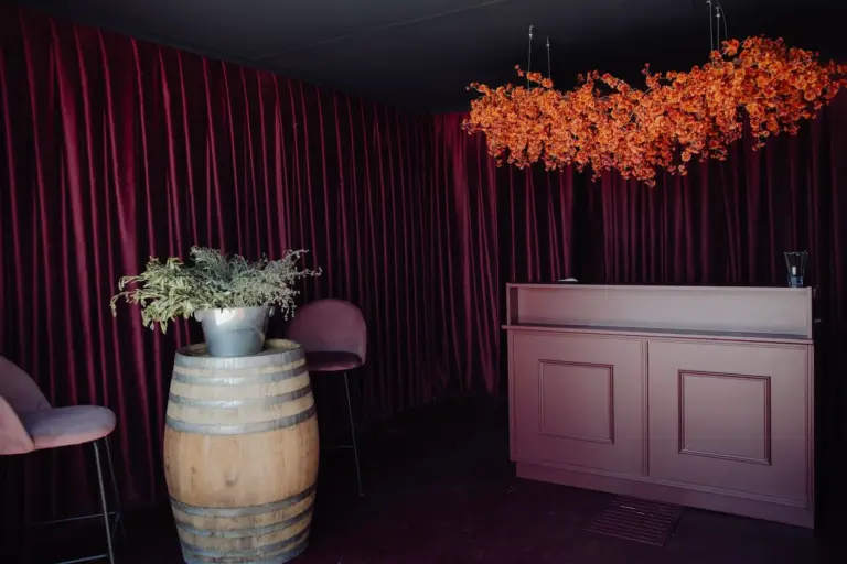 Ein schwach beleuchteter Raum mit burgunderroten Samtvorhängen, einem passenden Bartresen, einem Holzfass mit einer Topfpflanze, zwei Stühlen und einem Arrangement aus leuchtend orangefarbenen Blumen über dem Kopf.