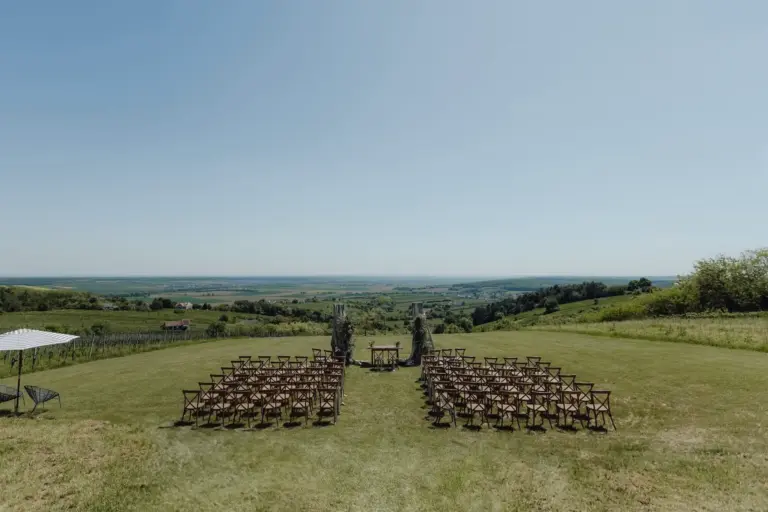 Leere Holzstühle stehen in Reihen auf einer Wiese für eine Hochzeitszeremonie, vor einem einfachen Torbogen mit Blick auf sanfte Hügel und einen klaren blauen Himmel im Hintergrund.