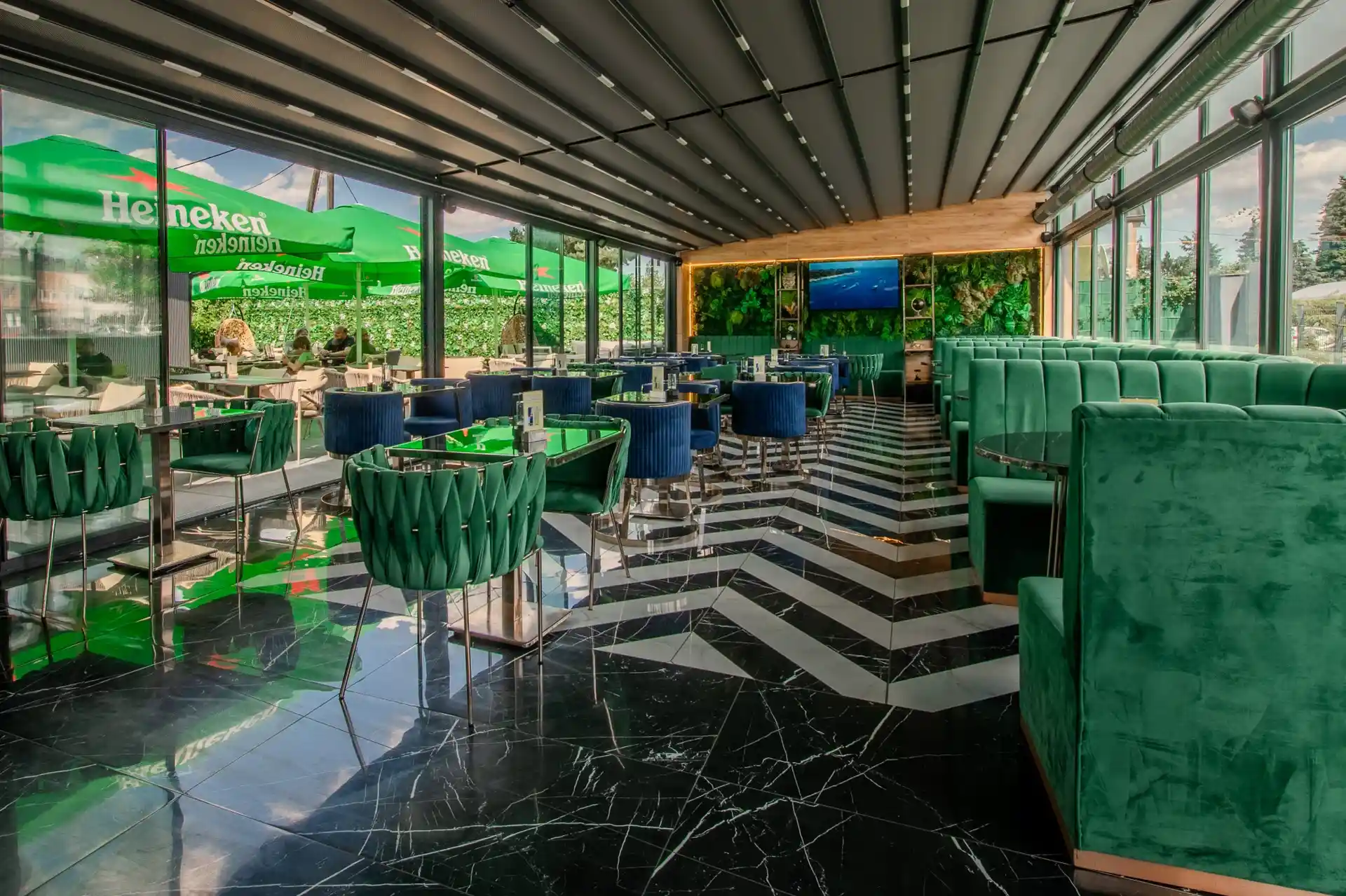 Modernes Restaurantinterieur mit grünen Samtstühlen, einem Boden mit Marmormuster, Glaswänden und einem durch die Fenster einsehbaren Außenbereich mit Heineken-Branding. Die Tische sind ordentlich angeordnet, und natürliches Licht durchflutet den Raum.