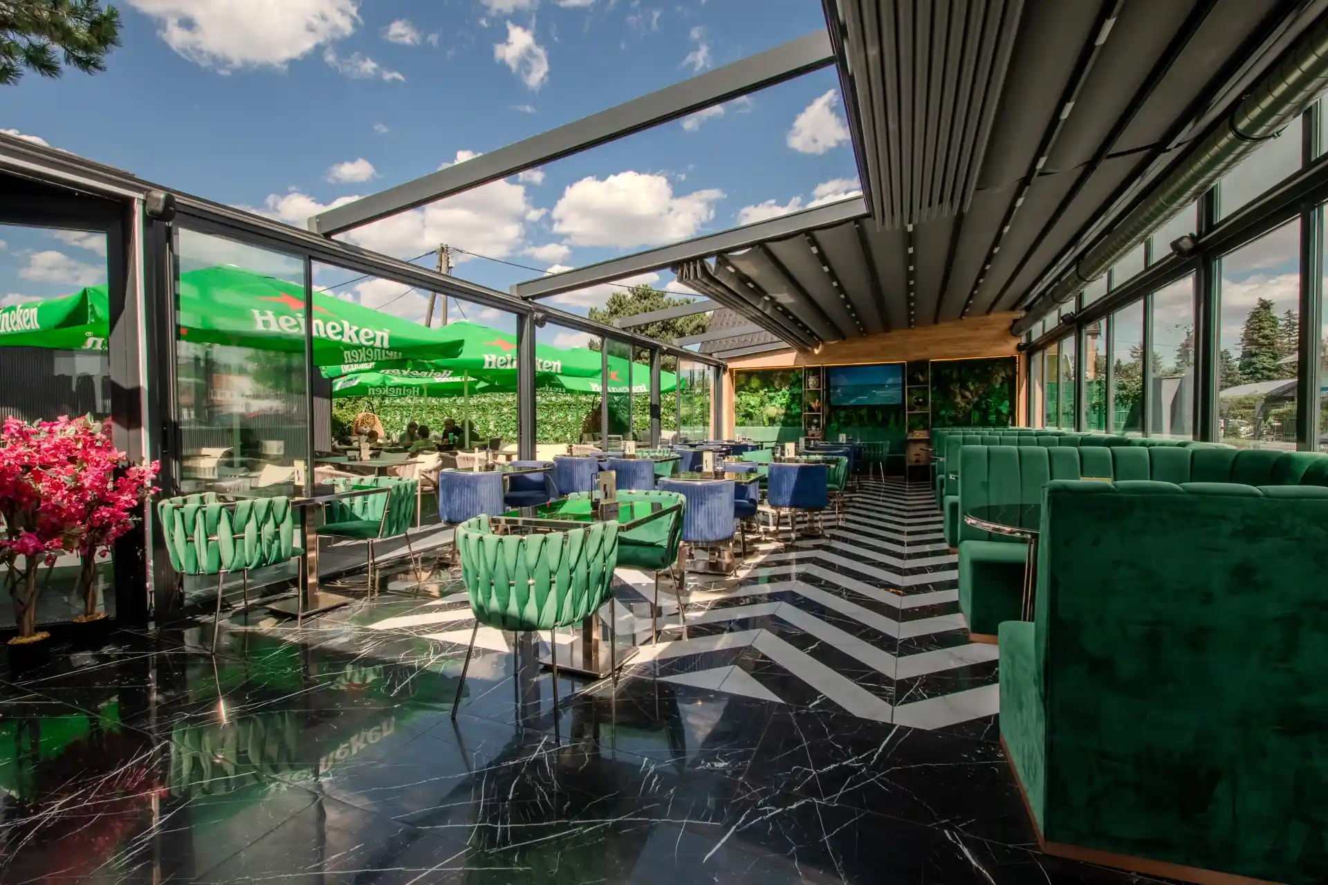 Chia Garden Modernes Restaurant mit Sitzgelegenheiten aus grünem und blauem Samt, Glaswänden, einem schwarzen Marmorboden mit weißem Zickzackmuster und einem teilweise offenen Dach. An sonnigen Tagen sind draußen grüne Sonnenschirme zu sehen.