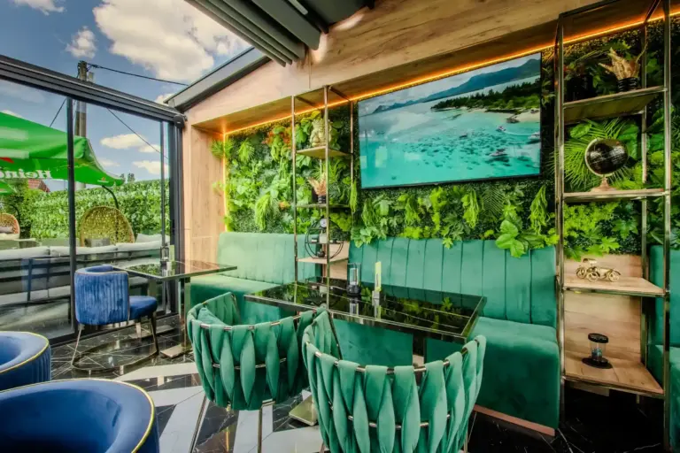 Chia Garden Schicke Lounge im Innenbereich mit grünen Samtsesseln, Glastischen, einer Wand aus tropischen Pflanzen, einem großen Strandfoto und einem durch die Glaswände sichtbaren Außenbereich unter blauem Himmel.