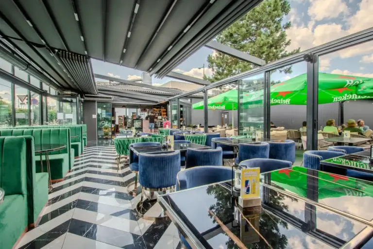 Ein modernes Café mit blauen Samtstühlen, glänzenden schwarzen Tischen, einem schwarz-weiß gestreiften Boden, Glaswänden und einem einfahrbaren Dach. Draußen sind grüne Sonnenschirme zu sehen, und der Raum wirkt hell und einladend.