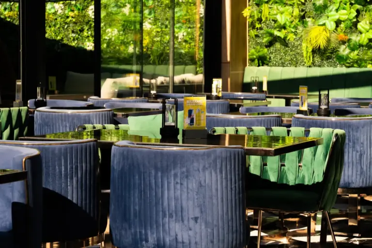 Ein modernes Restaurant mit blauen Samtstühlen an dunklen Tischen, großen Fenstern, in denen sich Bäume spiegeln, und einer üppig begrünten vertikalen Gartenwand. Das Sonnenlicht wirft Schatten und schafft so eine gemütliche, elegante Atmosphäre.