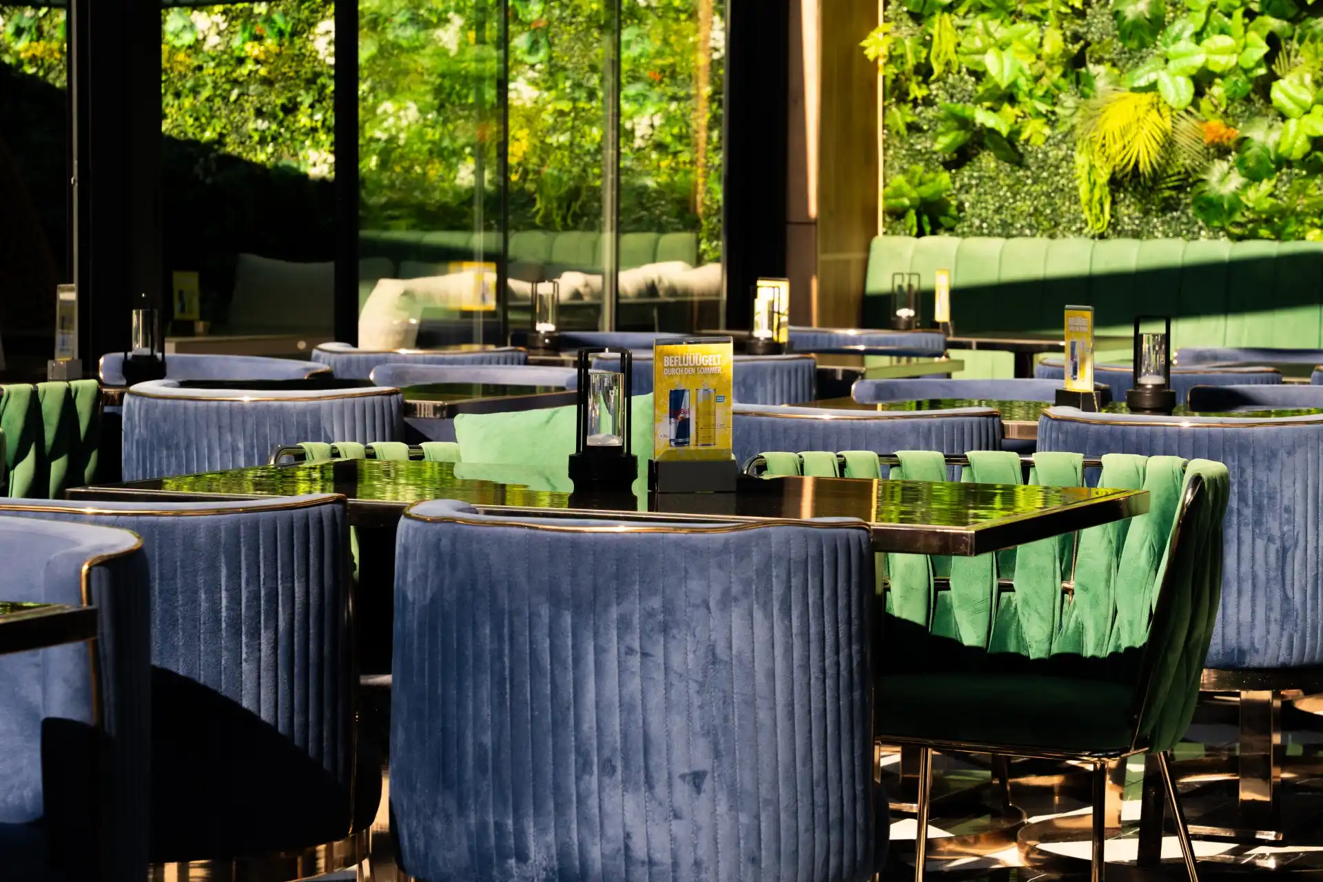 Ein modernes Restaurant mit blauen Samtstühlen an dunklen Tischen, großen Fenstern, in denen sich Bäume spiegeln, und einer üppig begrünten vertikalen Gartenwand. Das Sonnenlicht wirft Schatten und schafft so eine gemütliche, elegante Atmosphäre.