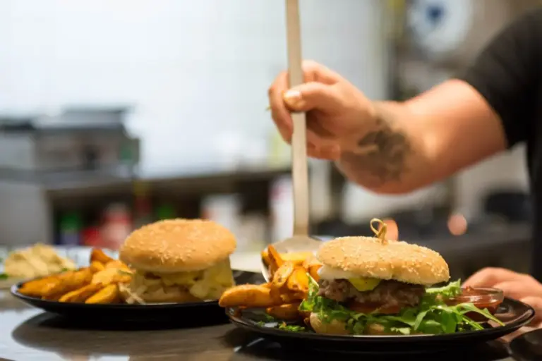 Eine Person serviert goldene Kartoffelspalten auf einem Teller mit einem Sesam-Burger, Salat und Tomate, neben einem weiteren Burger mit Sprossen, in der lebhaften Küche der Chill Out Lounge Wien.
