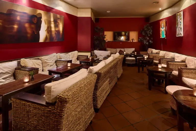 Gemütliche Lounge mit Korbsofas und -stühlen, dunklen Holztischen, roten Wänden, gedämpfter Beleuchtung und Kunstwerken an den Wänden, die eine warme und einladende Atmosphäre schaffen.