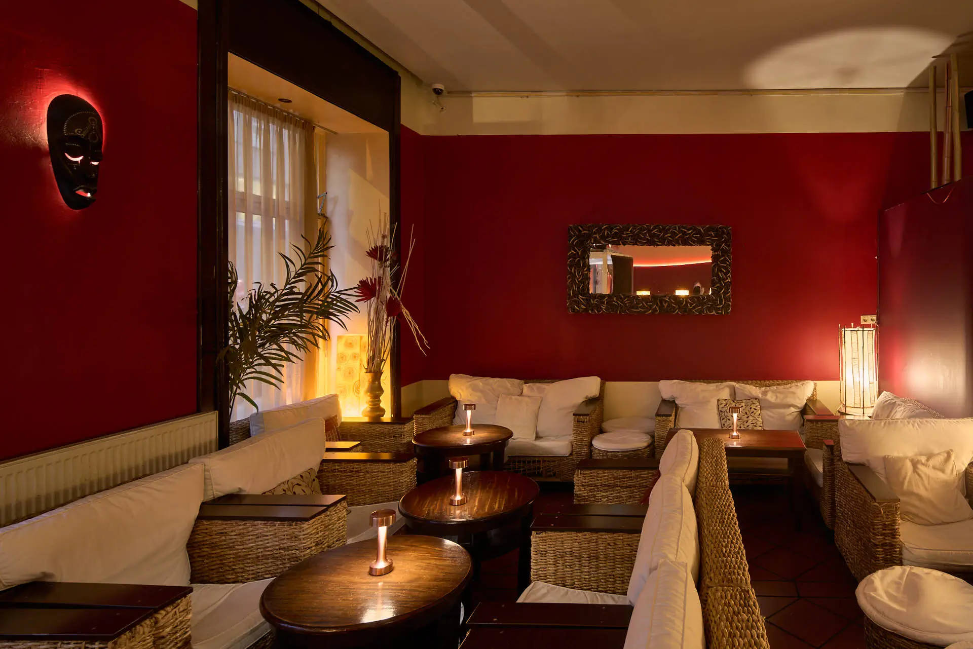 Eine gemütliche Lounge mit Korbsofas und weißen Kissen, warmes Licht von Tischkerzen und Lampen, rote Wände, ein großer Spiegel und dekorative Pflanzen schaffen eine entspannte und einladende Atmosphäre.