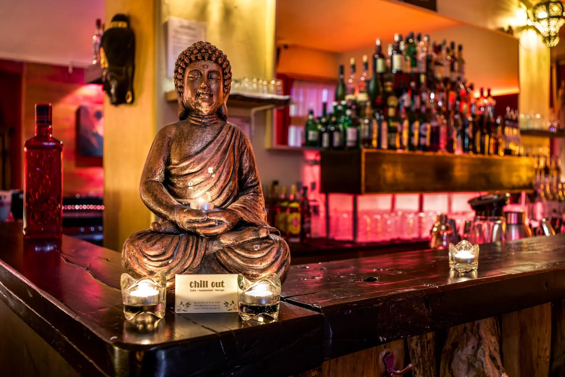 Eine bronzene Buddha-Statue mit einem Teelicht in der Hand steht auf einem hölzernen Bartresen, umgeben von kleinen Kerzen, während im schwach beleuchteten Hintergrund Alkoholflaschen und Gläser auf Regalen stehen.