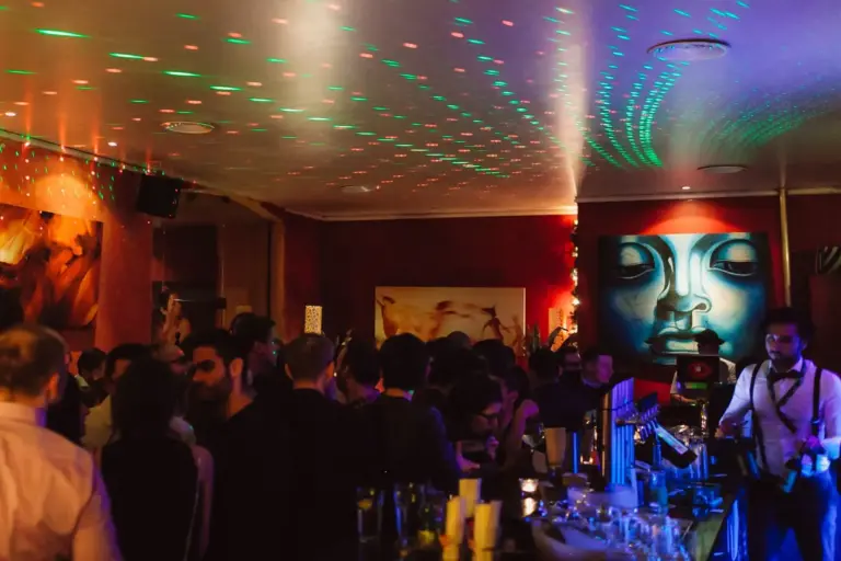 Eine überfüllte Bar mit Menschen, die sich unter bunten, gepunkteten Lichtern unterhalten. Die Wände sind mit lebendigen Kunstwerken geschmückt, darunter ein großes blaues Wandbild. Der Barkeeper serviert Getränke am Tresen.