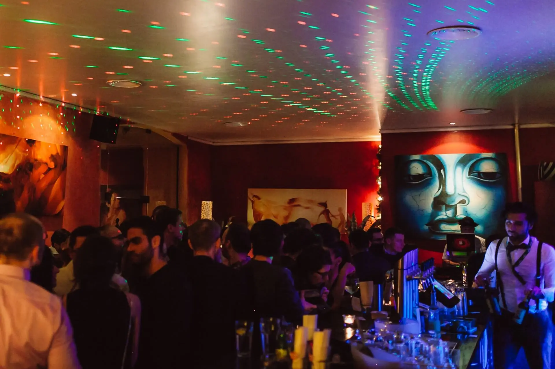 Eine überfüllte Bar mit Menschen, die sich unter bunten, gepunkteten Lichtern unterhalten. Die Wände sind mit lebendigen Kunstwerken geschmückt, darunter ein großes blaues Wandbild. Der Barkeeper serviert Getränke am Tresen.