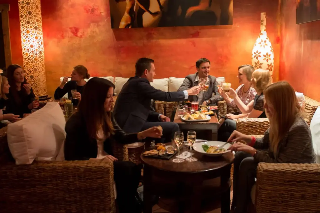 Eine Gruppe von Leuten sitzt auf Korbsofas um kleine Tische herum und genießt Getränke und Essen in einer gemütlichen, warm beleuchteten Lounge mit orange-roten Wänden und stimmungsvollem Dekor.