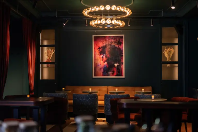 Eine stilvolle Lounge mit gedämpfter Beleuchtung, runden Kronleuchtern, einem hellbraunen Sofa, blauen Sesseln, kleinen Tischen mit Kerzenlicht und einem farbenfrohen abstrakten Gemälde an einer dunklen, blaugrünen Wand. Getrocknete Blumenarrangements stehen auf Regalen.