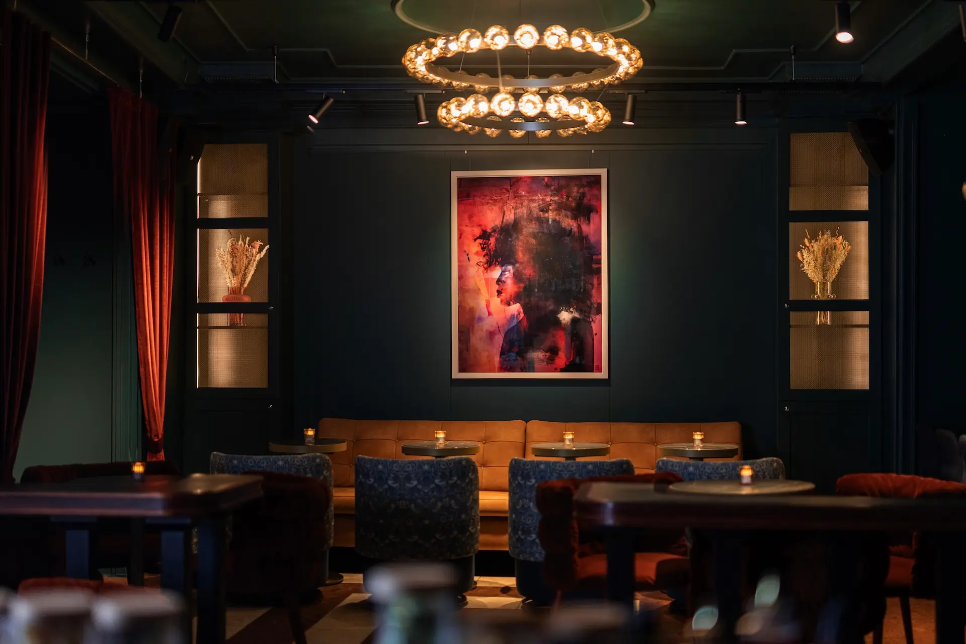 Eine stilvolle Lounge mit gedämpfter Beleuchtung, runden Kronleuchtern, einem hellbraunen Sofa, blauen Sesseln, kleinen Tischen mit Kerzenlicht und einem farbenfrohen abstrakten Gemälde an einer dunklen, blaugrünen Wand. Getrocknete Blumenarrangements stehen auf Regalen.