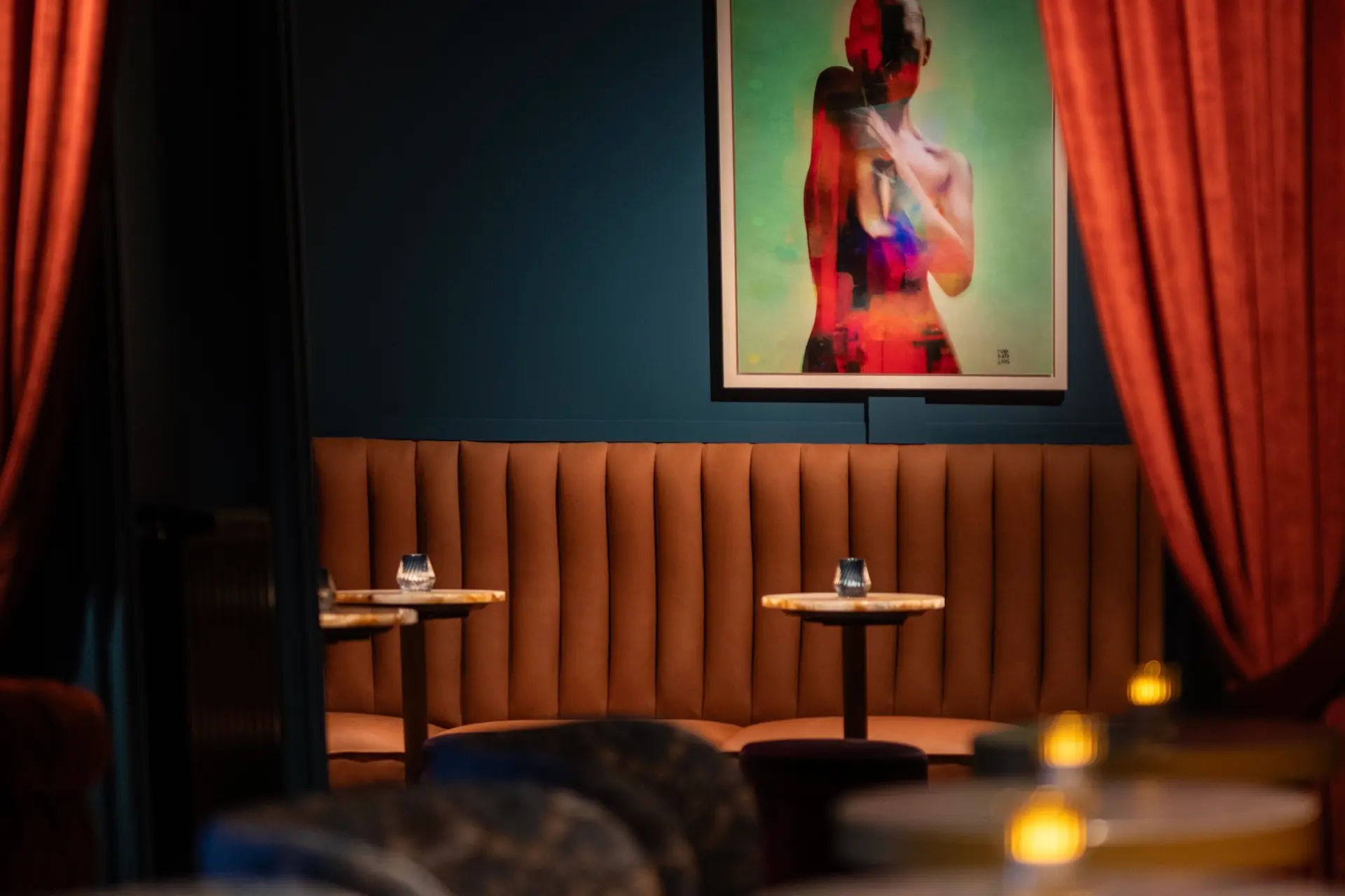 Eine schwach beleuchtete Lounge mit hellbraunen gepolsterten Sitzmöbeln, kleinen runden Tischen, Kerzen, roten Vorhängen und einem bunten abstrakten Kunstwerk einer Person an der blauen Wand.