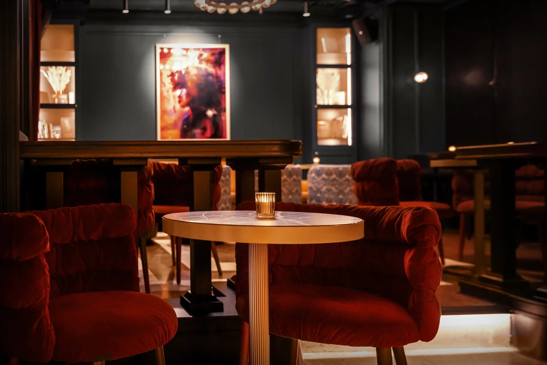 Eine gemütliche Lounge mit roten Plüschsesseln um einen runden Holztisch mit einer brennenden Kerze. Der Raum ist schummrig beleuchtet, mit abstrakter Kunst und Fenstern im Hintergrund, die eine intime Atmosphäre schaffen.