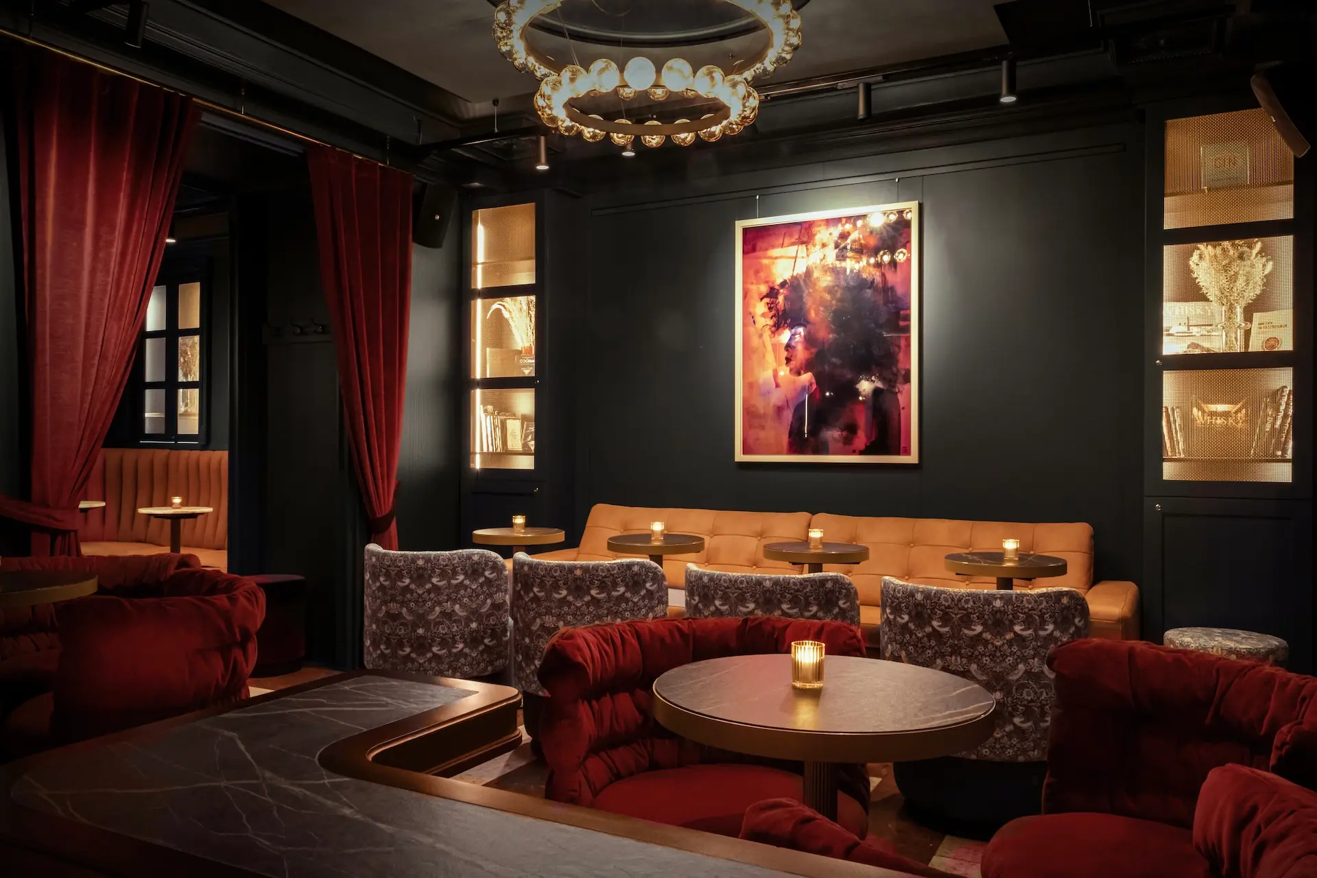 Eine schwach beleuchtete Lounge mit roten Plüschsesseln, einem hellbraunen Sofa, runden Tischen mit Kerzen, dunklen Wänden, Samtvorhängen und einem gerahmten abstrakten Kunstwerk, das über dem Sitzbereich beleuchtet wird.