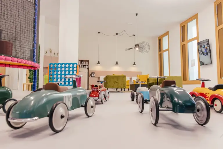 Ein helles Spielzimmer mit mehreren Spielzeug-Tretautos im Vintage-Stil auf dem Boden, bunten Möbeln, großen Fenstern und verspielter Dekoration im Hintergrund.